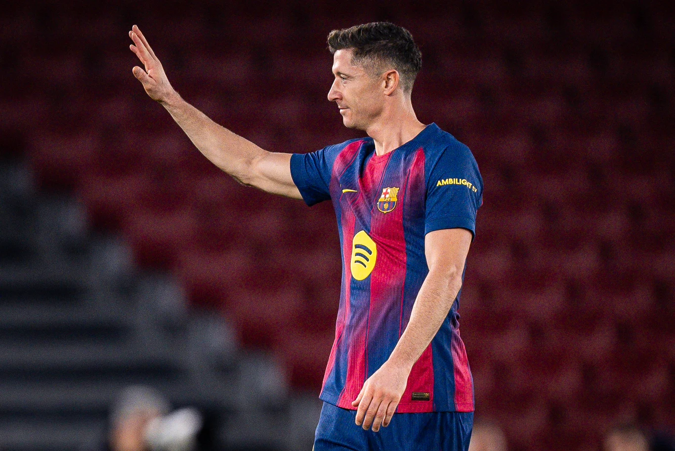 Piłkarz w stroju FC Barcelony stoi na boisku, unosząc prawą rękę w geście pozdrowienia lub pożegnania. W tle rozmyte trybuny stadionu.