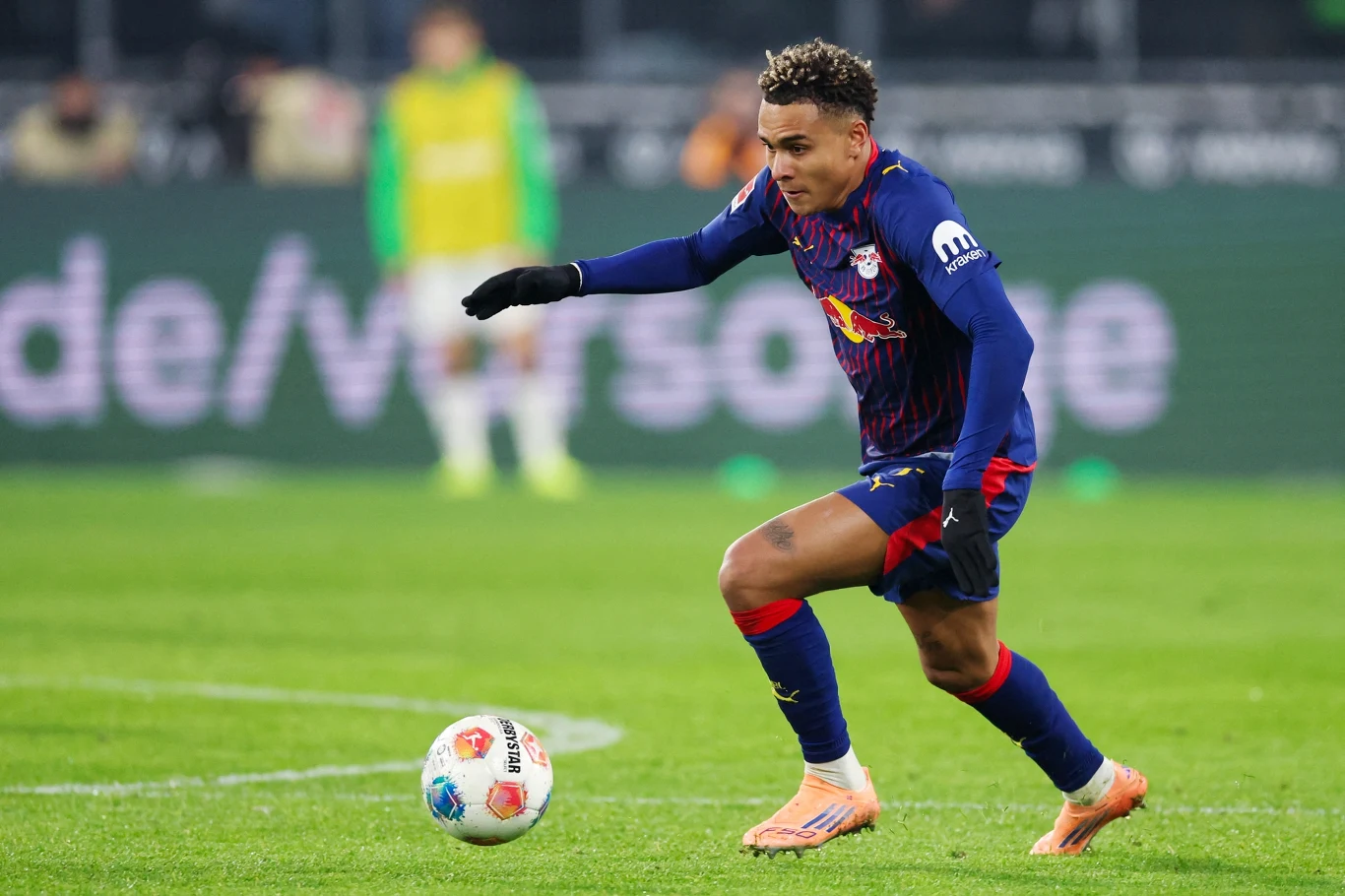 Piłkarz w niebieskim stroju z logo klubu RB Lipsk biegnący z piłką po murawie podczas meczu piłki nożnej, w tle rozmyte sylwetki innych zawodników oraz elementy stadionu.