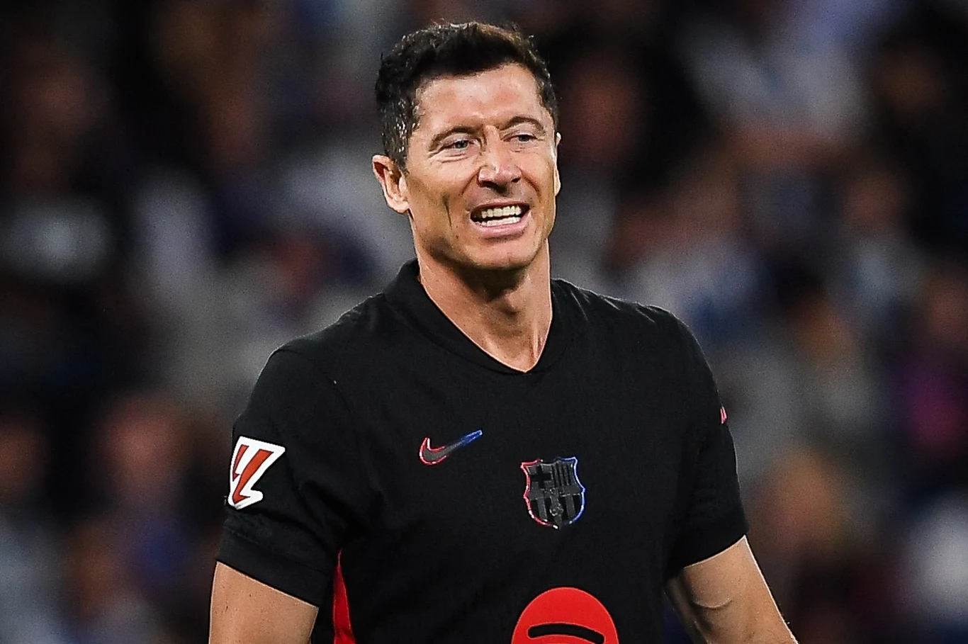 Robert Lewandowski Piłkarz w czarnej koszulce z herbem klubu FC Barcelona i czerwonym logo sponsora na tle niewyraźnej publiczności podczas meczu.