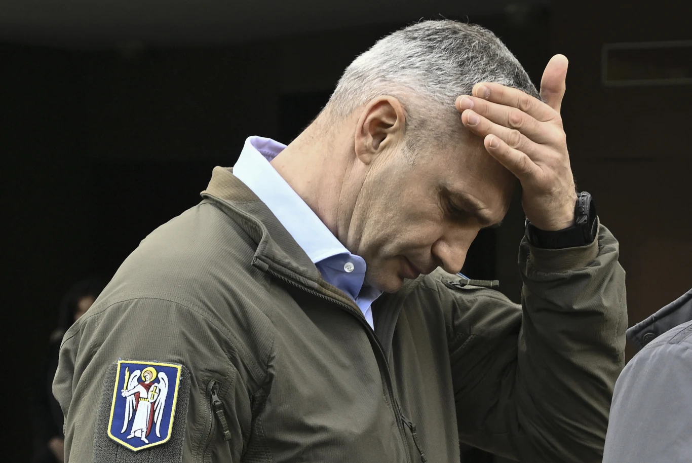 Mężczyzna o siwych włosach w zielonej kurtce dotyka czoła dłonią w geście zamyślenia lub zmartwienia. Na ramieniu widoczna naszywka z herbem przedstawiającym anioła.