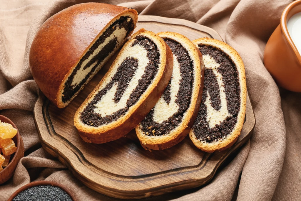 Makowiec tradycyjny Trzy kromki tradycyjnego makowca o wyraźnej spirali makowej na przekroju, ułożone na drewnianej desce, obok całego bochenka. W tle lniana serwetka i naczynie z mlekiem.