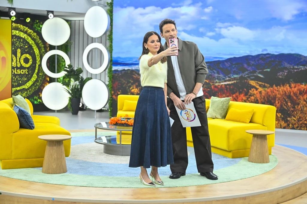 Dwoje prezenterów na tle kolorowego, nowoczesnego studia telewizyjnego, robiących wspólne selfie. Wnętrze utrzymane w jasnych barwach z żółtą sofą, dekoracyjnymi okręgami na ścianie i dużym ekranem przedstawiającym pejzaż górski.