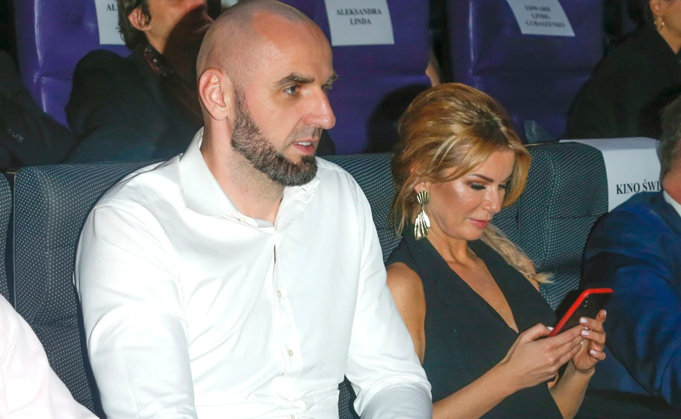 Marcin Gortat z żoną Żanetą Gortat-Stanisławską Dwójka elegancko ubranych osób siedzi obok siebie w fotelach na sali, mężczyzna w białej koszuli patrzy przed siebie, a kobieta w czarnej sukni przegląda telefon.