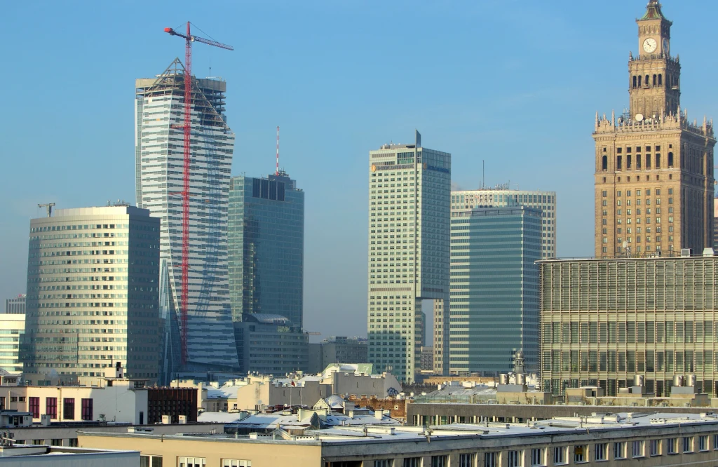Panorama centrum Warszawy z charakterystycznymi nowoczesnymi wieżowcami, dźwigiem budowlanym oraz Pałacem Kultury i Nauki po prawej stronie kadru, na pierwszym planie dachy niższych budynków.