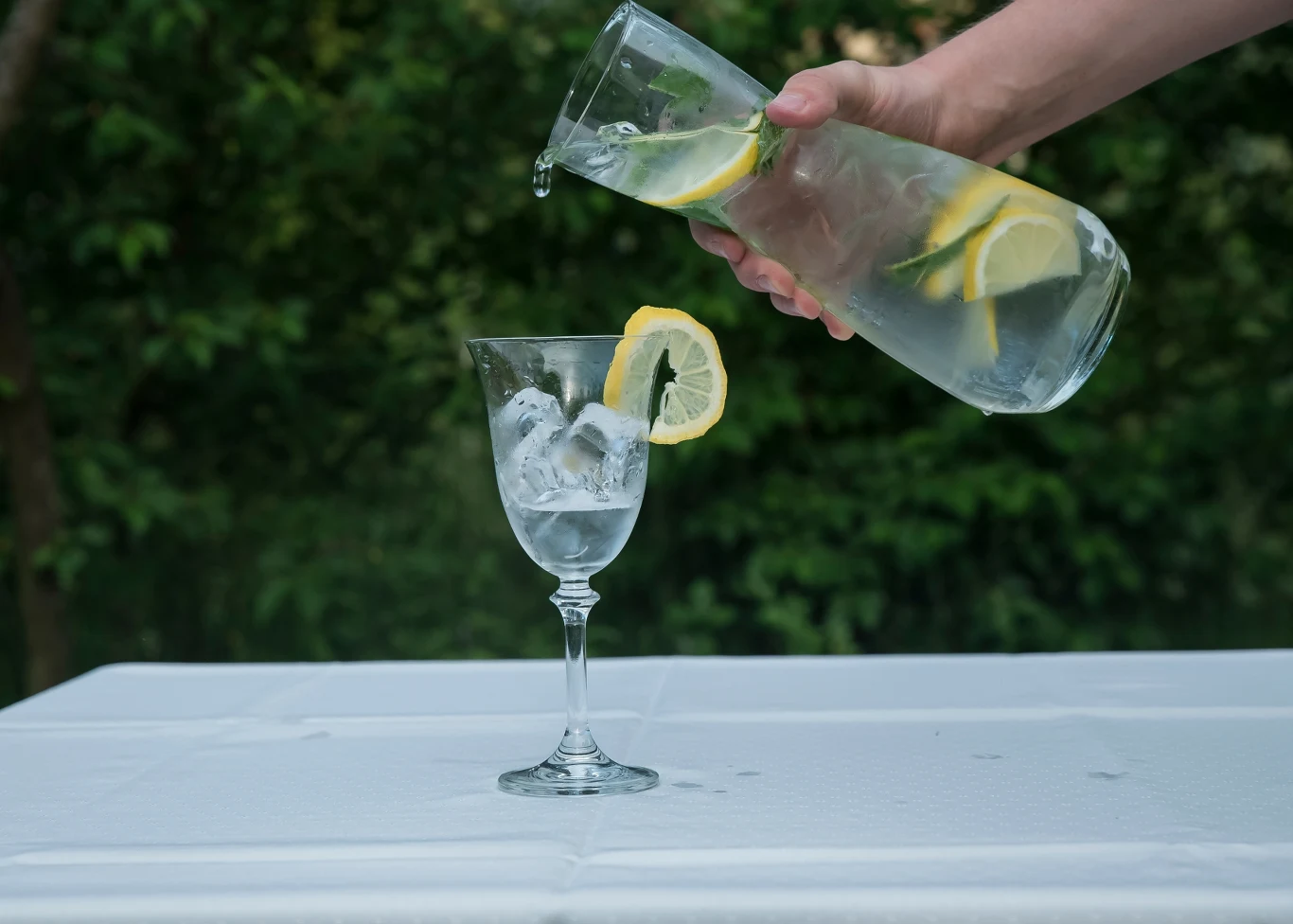 Dlaczego latem musisz uważać na odwodnienie? Szklanka z lodem i plasterkiem cytryny na brzegu, do której nalewana jest woda z dzbanka z plasterkami cytryny i limonki, całość stoi na białym stole w ogrodowym otoczeniu.
