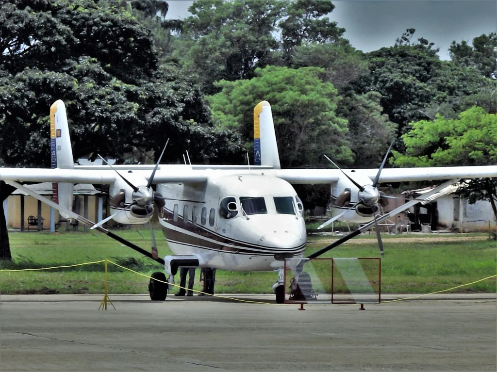 PZL M28 Skytrucker Gwardii Narodowej Wenezueli Dwusilnikowy samolot pasażerski z napędem śmigłowym stoi zaparkowany na płycie lotniska, otoczony żółtymi barierkami zabezpieczającymi, w tle widoczne zabudowania i gęste zielone drzewa.