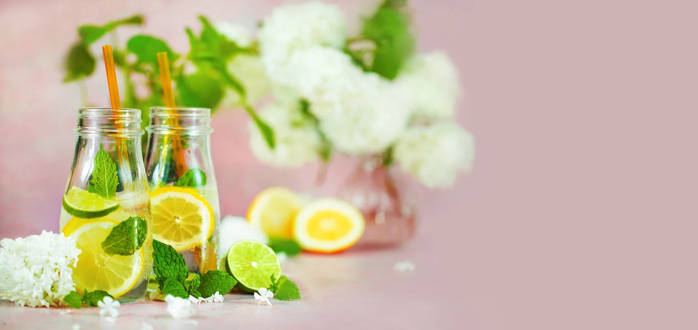 Smak i jakość wody mają znaczenie Dwie butelki z napojem cytrynowym z plasterkami cytryny, limonki oraz świeżą miętą, ozdobione kolorowymi słomkami. W tle jasne kwiaty, przekrojona cytryna i delikatnie rozmyty różowy gradient.