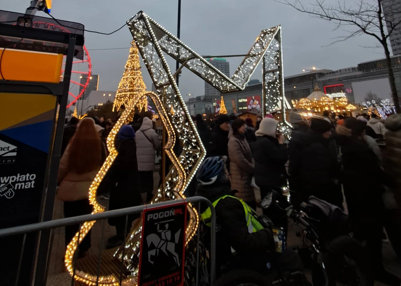Wejście na jarmark w Warszawie. Już na początku zaczyna się "kolejka" Tłum osób zebranych wokół świątecznych dekoracji z motywami świetlnymi, w tle widoczna choinka i podświetlana litera M, atmosfera zimowa z miejską infrastrukturą w tle.