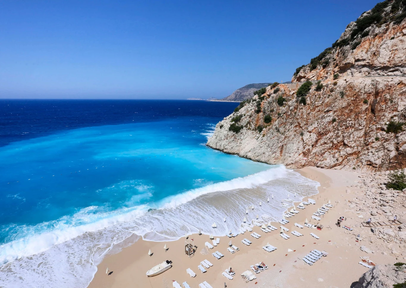 Wakacje Turcja 2026 z Coral Travel Piaszczysta plaża z białymi leżakami i parasolami, otoczona przez masywną skalistą ścianę, lazurowa woda morza łagodnie załamuje się o brzeg pod bezchmurnym, niebieskim niebem.