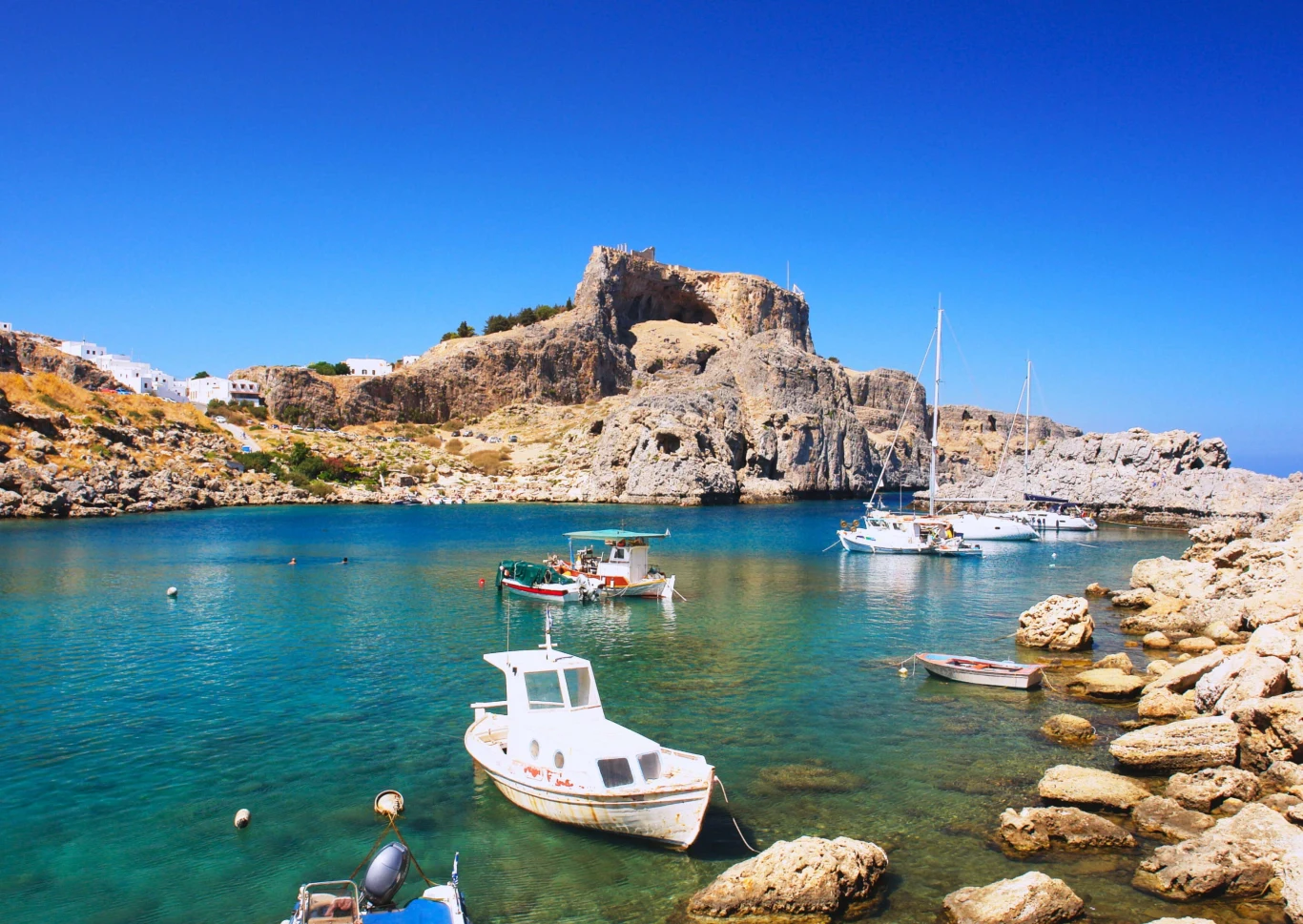 Wakacje Grecja 2026 z Coral Travel Mała marina z białymi łódkami na krystalicznie czystej wodzie, w tle skaliste wzgórze z ruinami starożytnej twierdzy; spokojna, jasna pogoda i błękitne niebo.