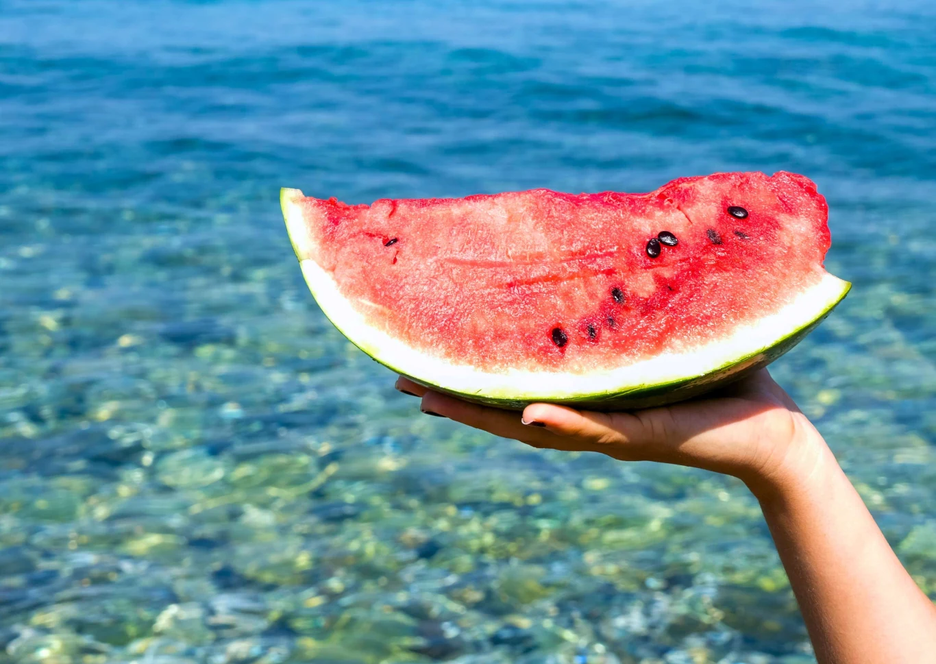 Gdzie taniej na wakacje 2026 z Coral Travel – Turcja czy Grecja? Duży kawałek arbuza trzymany w dłoni na tle czystej, błękitnej wody.