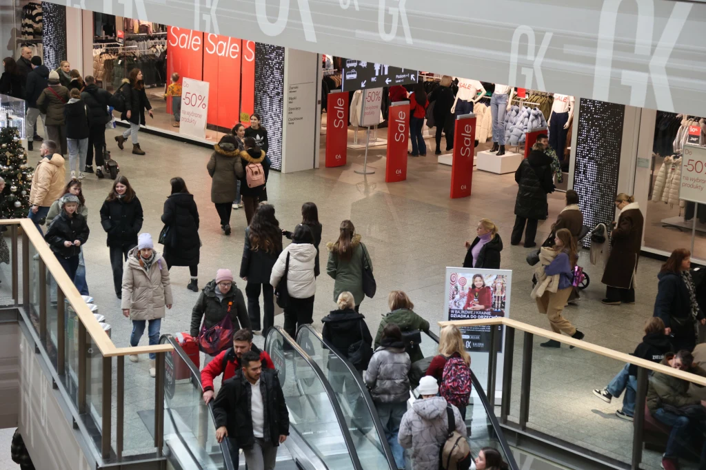 Tłum ludzi poruszający się po galerii handlowej. W tle liczne witryny sklepowe z widocznymi napisami promocyjnymi oraz tablicami informującymi o wyprzedażach. Na pierwszym planie osoby korzystają z ruchomych schodów.