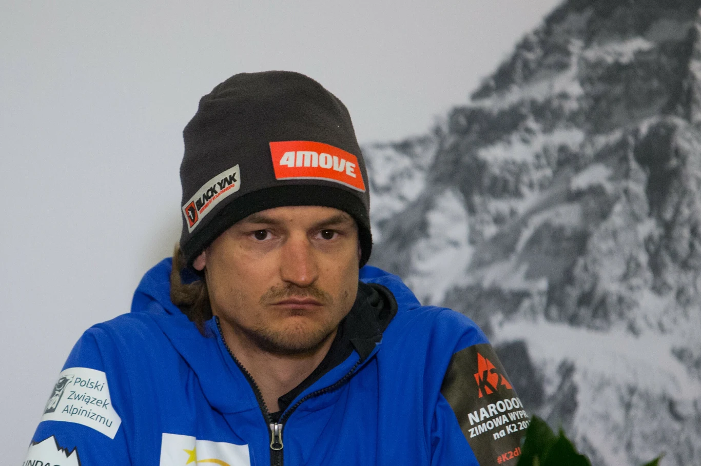 Adam Bielecki Mężczyzna w niebieskiej kurtce z naszywkami oraz czapce z logo, siedzi na tle czarno-białego zdjęcia górskiego krajobrazu, patrzy poważnie przed siebie.