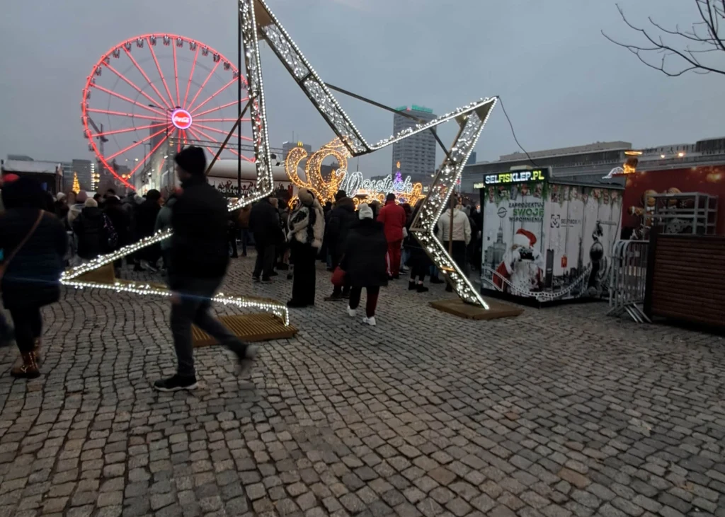 Ruchliwy jarmark świąteczny po zmroku, ozdobiony dużą świetlną gwiazdą i świątecznymi dekoracjami, w tle widoczne diabelskie koło oraz liczne stoiska i grupy ludzi.