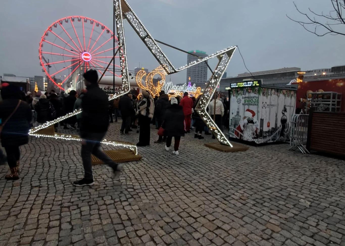 Świątecznych dekoracji na jarmarku jest niewiele Ruchliwy jarmark świąteczny po zmroku, ozdobiony dużą świetlną gwiazdą i świątecznymi dekoracjami, w tle widoczne diabelskie koło oraz liczne stoiska i grupy ludzi.