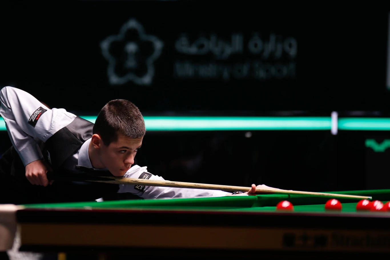 Michał Szubarczyk Młody zawodnik koncentruje się nad precyzyjnym uderzeniem bilą podczas meczu snookera. Elegancki strój turniejowy, profesjonalny stół z czerwoną bilą w skupieniu sportowca oraz ciemne tło nadające scenie powagę i dramaturgię.