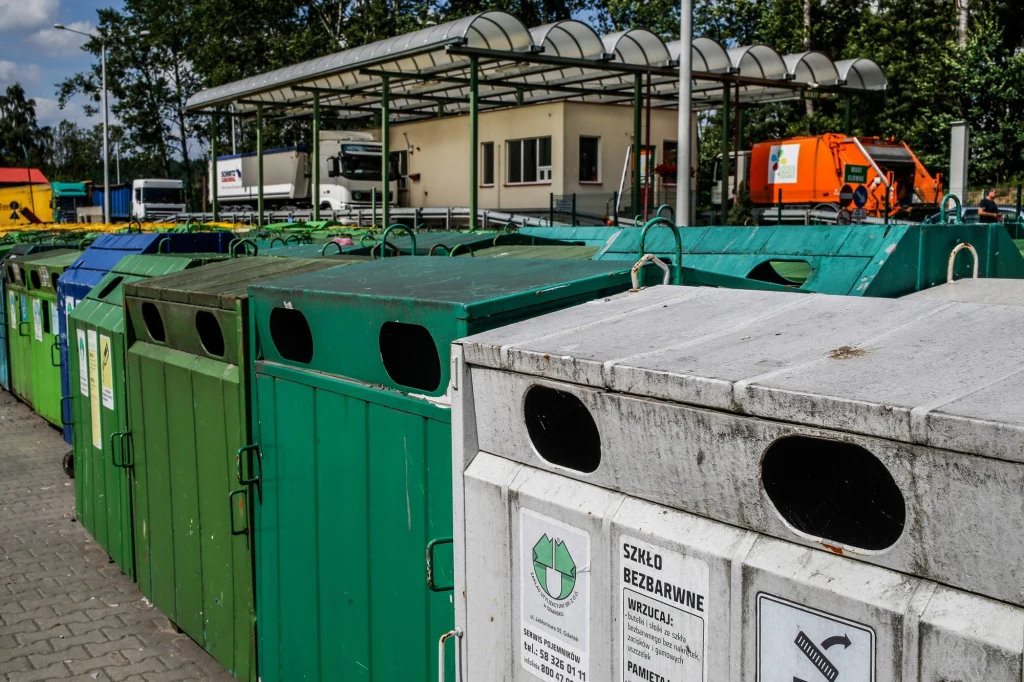 Rząd dużych pojemników na odpady segregowane w różnych kolorach ustawionych na placu, w tle widoczna wiata oraz budynek administracyjny i pojazdy związane z gospodarką odpadami.
