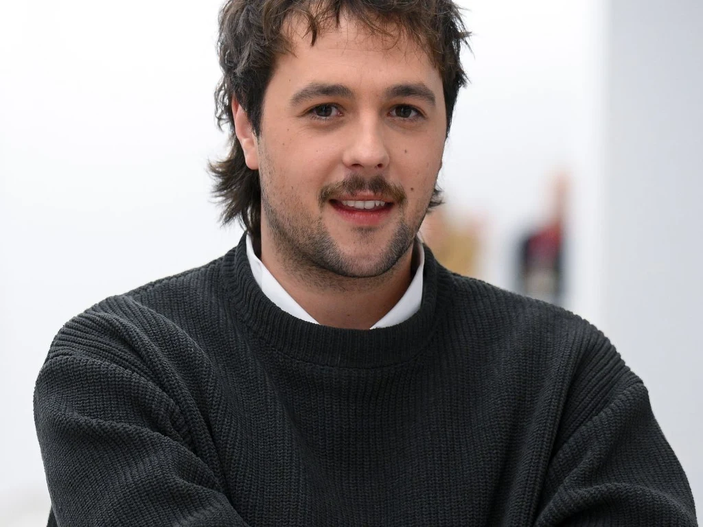 Jan Piwowarczyk Młody mężczyzna z ciemnymi włosami i zarostem uśmiecha się delikatnie, ubrany w ciemny sweter na białą koszulę, stoi na tle jasnego, rozmytego pomieszczenia.