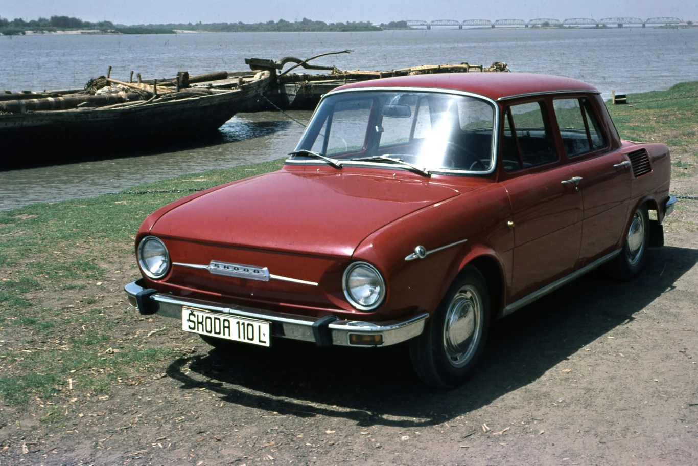 Czerwony samochód osobowy marki Škoda 110 L zaparkowany nad brzegiem rzeki, w tle drewniana łódź oraz most na horyzoncie.