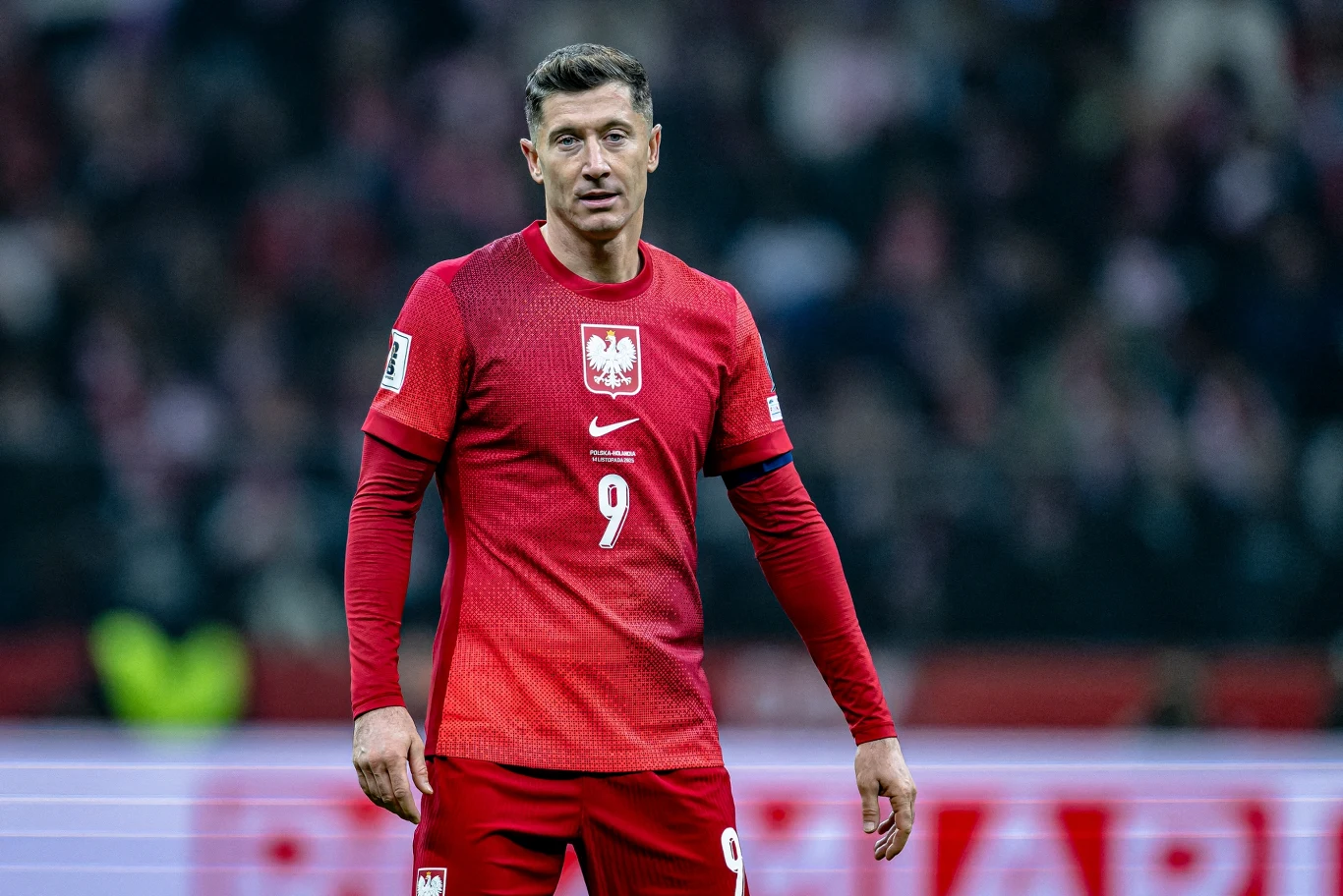 Piłkarz w czerwonym stroju reprezentacji Polski z numerem 9 stoi na murawie stadionu podczas meczu piłki nożnej, w tle widoczni kibice oraz zamazana trybuna.