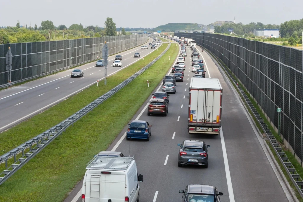 Korek samochodowy na autostradzie, liczne pojazdy w jednym kierunku stoją w zatorze, podczas gdy przeciwległa nitka drogi jest niemal pusta. Wzdłuż autostrady ustawione są ekrany dźwiękochłonne, wokół znajdują się tereny zielone.