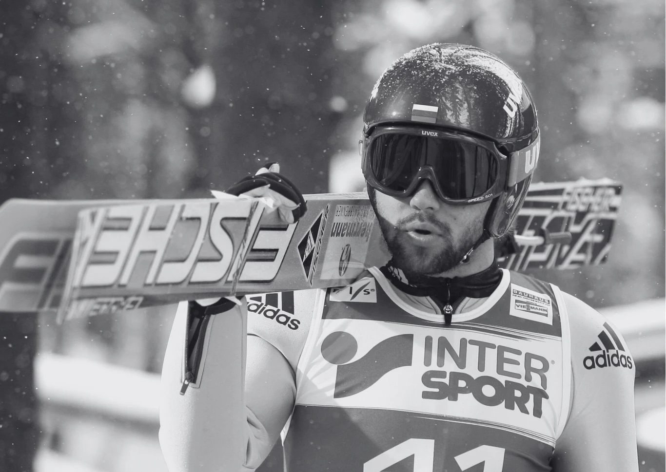 Paweł Karelin Narciarz w sportowym stroju i kasku trzyma narty na ramieniu, lekko otwarte usta, widoczne logo Intersport i Adidas na stroju, śnieżny krajobraz w tle, ujęcie w czerni i bieli.