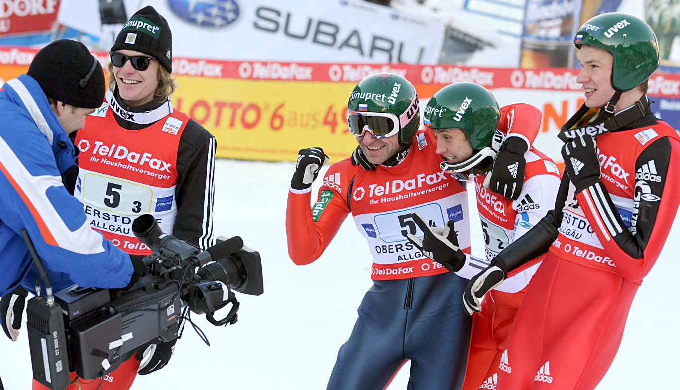 Paweł Karelin wraz z kadrą rosyjskich skoczków podczas konkursu drużynowego na mistrzostwach świata w 2009 roku Czterech sportowców ubranych w kombinezony narciarskie i kaski pozuje uśmiechając się przed kamerą telewizyjną na zawodach zimowych, w tle widoczne reklamy sponsorów na śniegu.