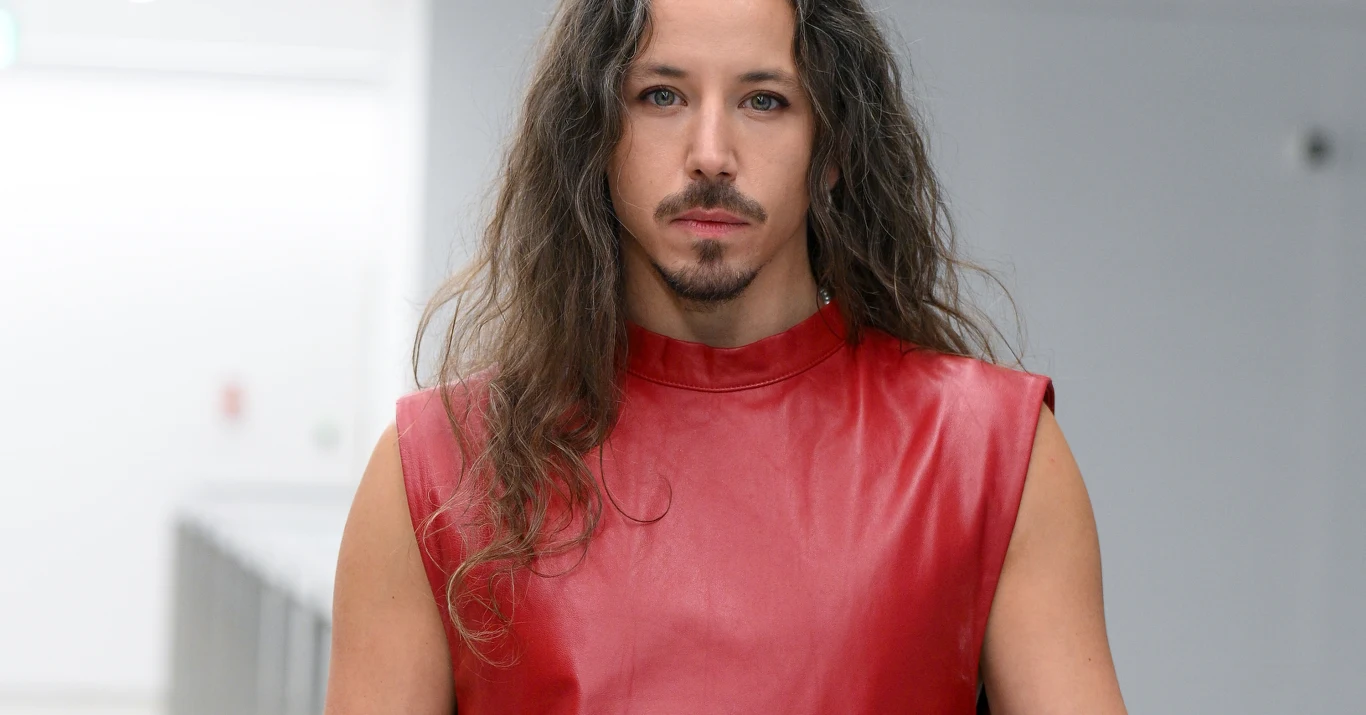 Michał Szpak Mężczyzna o długich, kręconych włosach i lekkim zaroście ubrany w czerwoną, bezrękawną bluzkę na tle jasnego, nowoczesnego wnętrza.