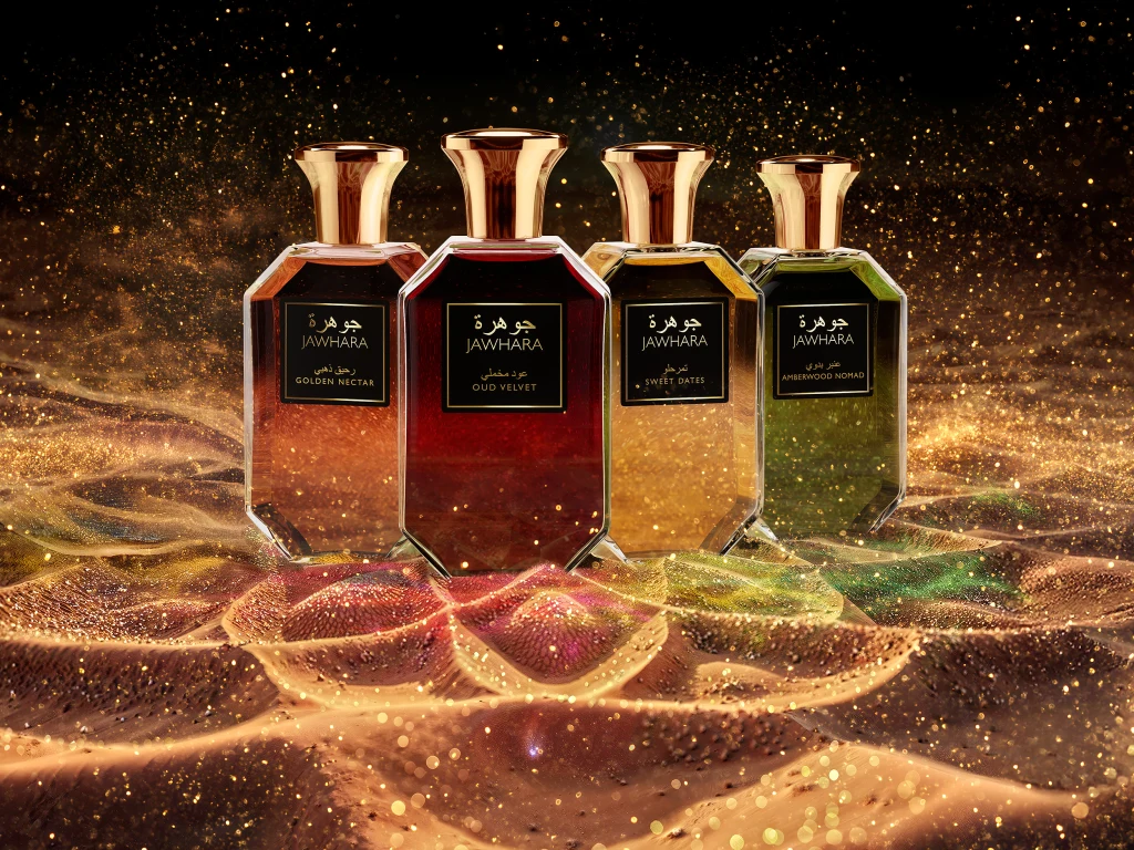Cztery eleganckie flakony perfum marki Jawhara ustawione w szeregu na błyszczącym, złocistym tle przypominającym rozsypany brokat lub piasek, każdy flakon ma złotą nakrętkę i etykietę z nazwą zapachu.