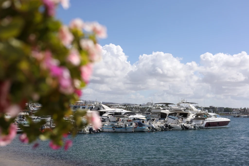Luksusowe jachty zacumowane w marinie na tle błękitnej wody i nieba z białymi chmurami, na pierwszym planie rozmyte różowe kwiaty.