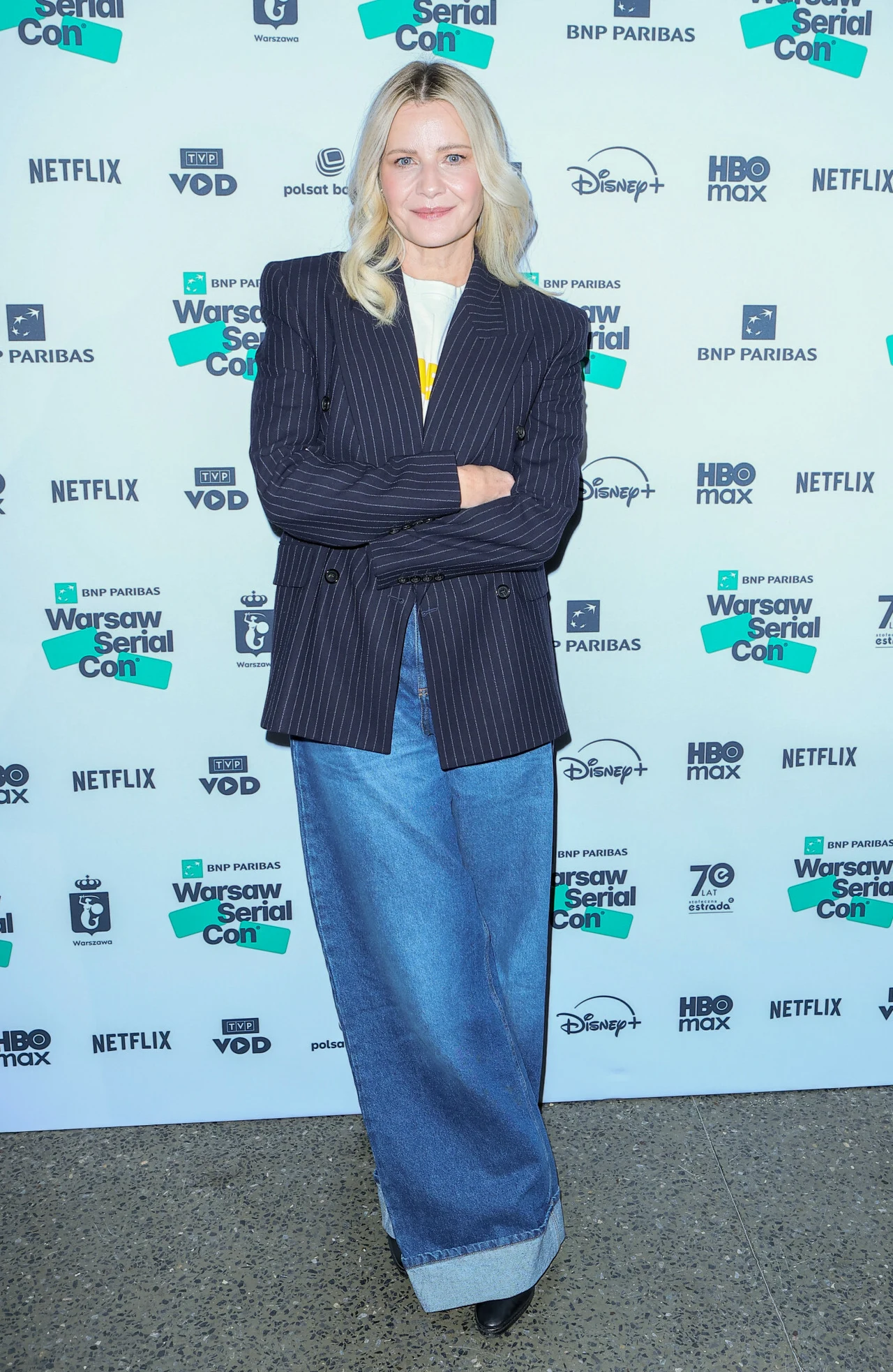 Kobieta o blond włosach stoi na tle ścianki z logotypami różnych firm i organizacji, ubrana w ciemną marynarkę w prążki i szerokie, niebieskie dżinsy. Pozuje z ramionami skrzyżowanymi i uśmiechem na twarzy.