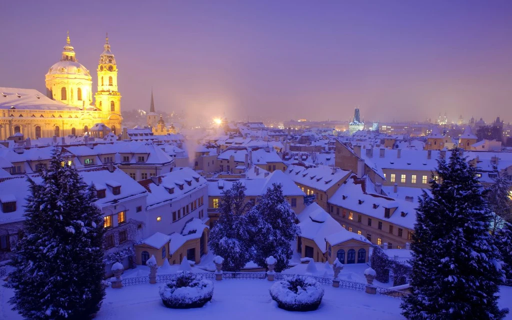 Zimowy krajobraz miasta z licznymi ośnieżonymi dachami, zabytkowy kościół z jasnym oświetleniem po lewej stronie, fioletowo-niebieskie niebo, drzewa pokryte śniegiem i uspokajająca atmosfera wieczorna.