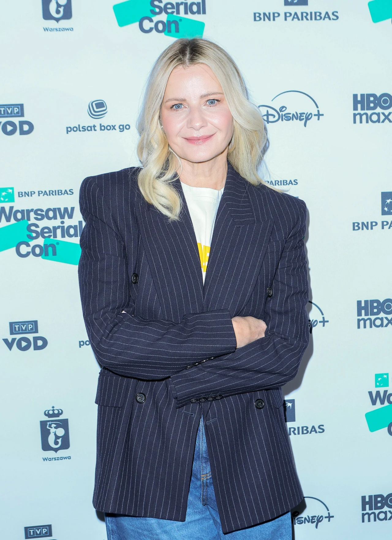 Kobieta z blond włosami ubrana w granatową marynarkę w prążki, jasny T-shirt oraz jeansy, stoi na tle ścianki z logotypami różnych marek i wydarzenia Warsaw Serial Con, prezentując zrelaksowaną postawę z założonymi rękami.