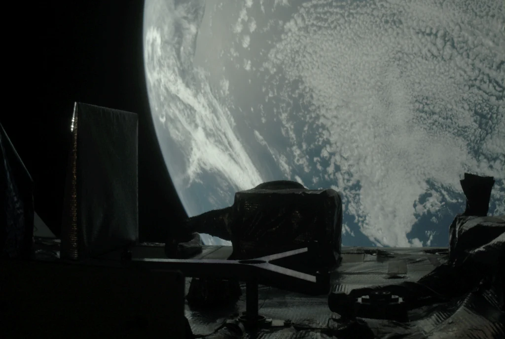 Widok Ziemi z kosmosu, na pierwszym planie fragmenty satelity lub sondy kosmicznej pokryte metalowymi i foliowymi materiałami, w tle widać powierzchnię planety pokrytą chmurami.