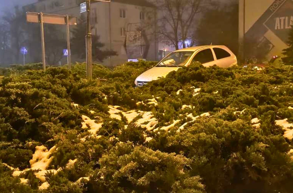 Samochód osobowy utknął w gęstych krzewach na środku ronda, wokół widoczne są resztki śniegu oraz budynki mieszkalne w tle.