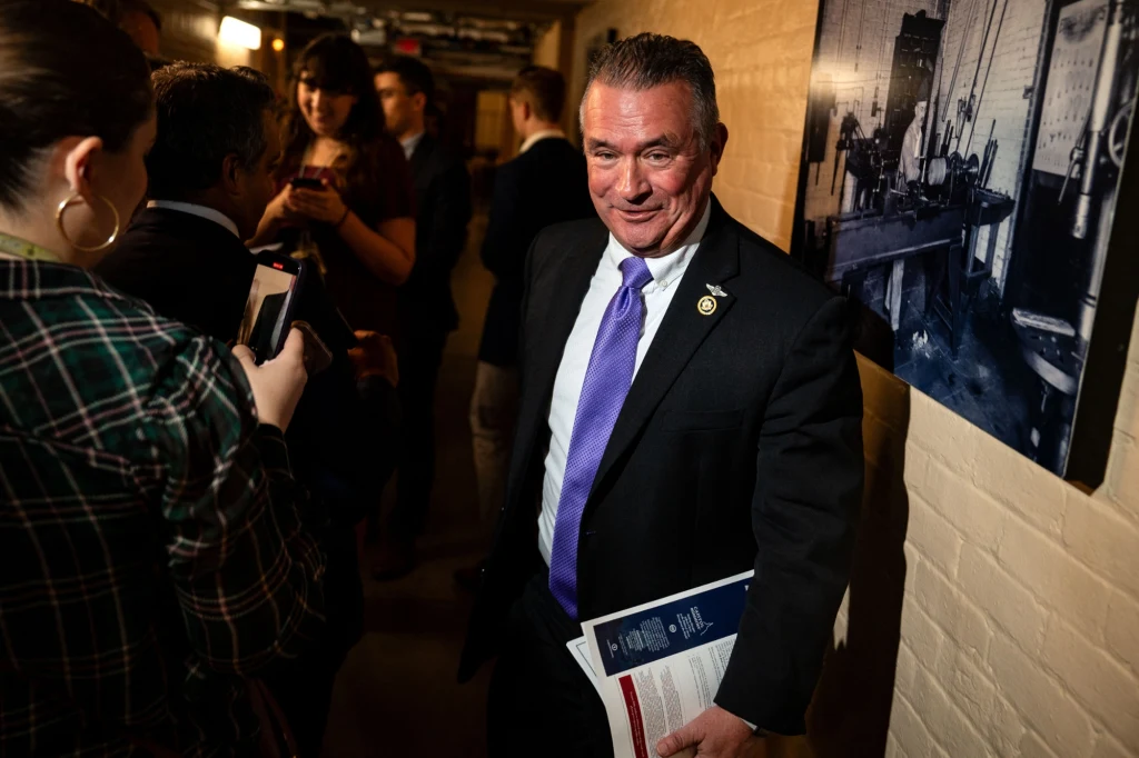 Mężczyzna w garniturze z przypiętą odznaką stoi w korytarzu oświetlonym ciepłym światłem, trzymając dokumenty. W tle widoczna grupa ludzi, którzy rozmawiają i korzystają z telefonów, na ścianie zawieszono czarno-białą fotografię industrialną.