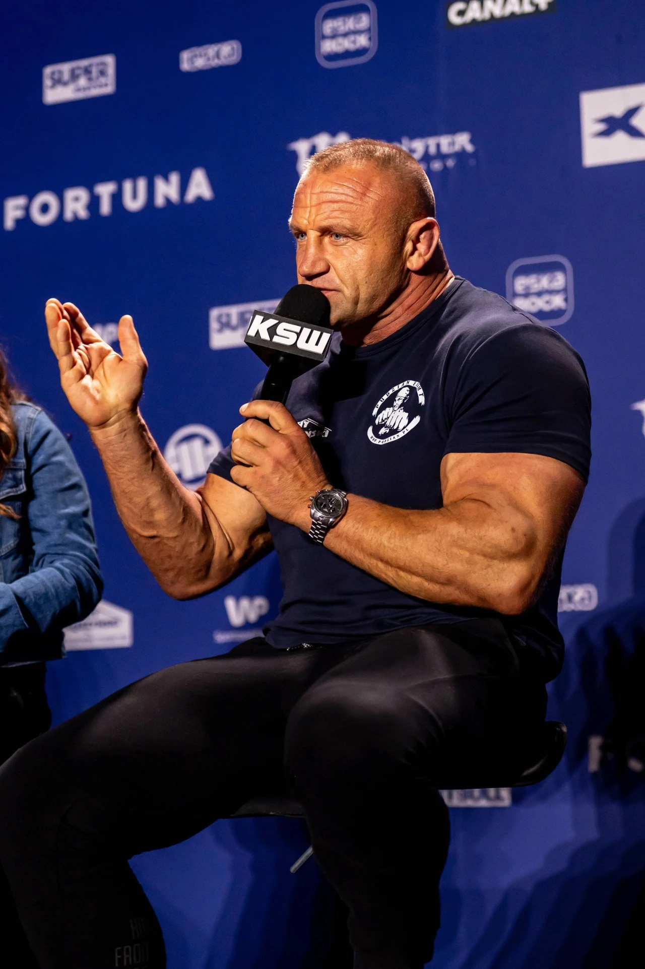 Umięśniony mężczyzna w koszulce sportowej siedzi na krześle i trzyma mikrofon z logo KSW, gestykuluje podczas konferencji prasowej, w tle niebieska ściana z logotypami sponsorów.