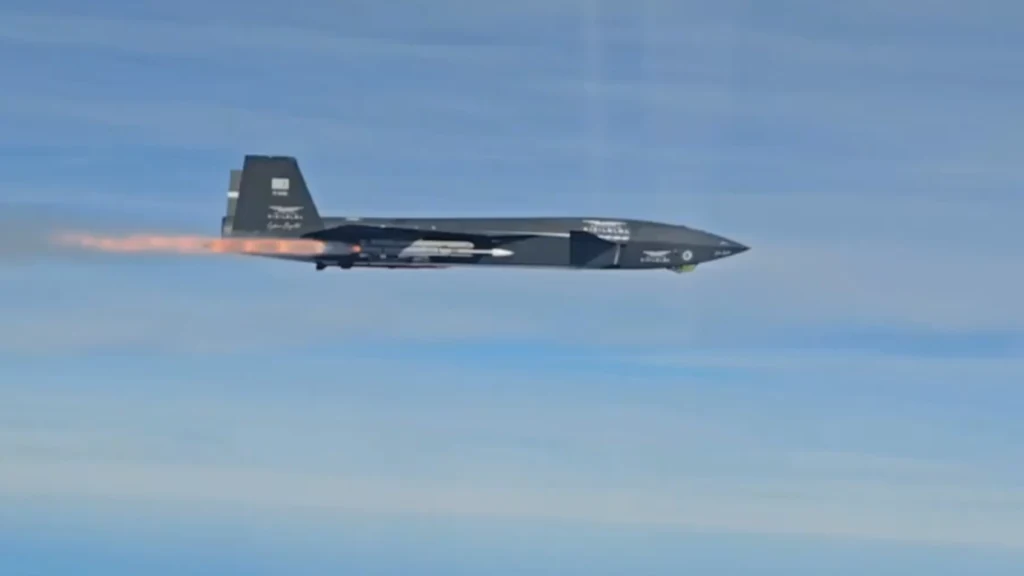 Czarny wojskowy samolot odrzutowy z widocznym ogniem wydobywającym się z silnika podczas lotu na tle jasnego nieba.