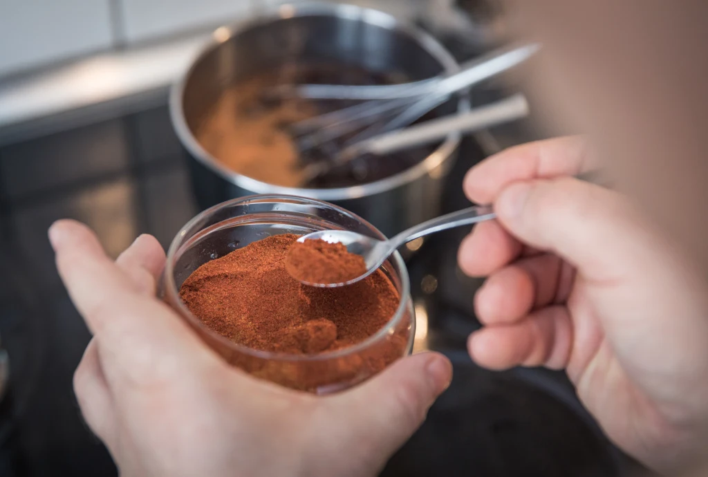 Ręce trzymają słoik z czerwoną przyprawą oraz łyżeczkę nabierającą jej porcję. W tle na kuchence stoi garnek z gotującą się potrawą oraz narzędzia kuchenne.