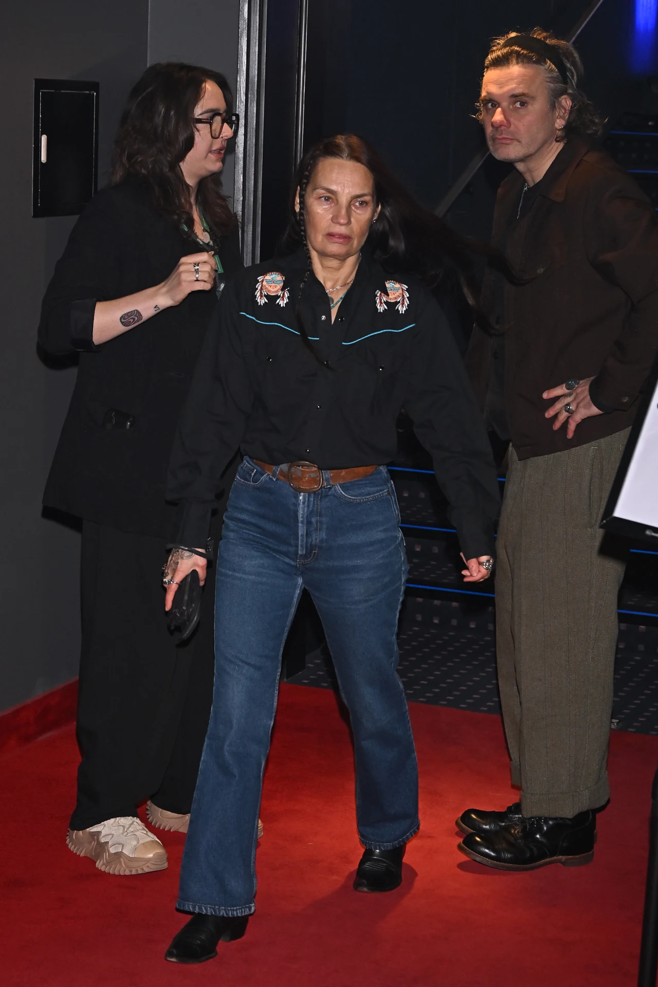 Trzy osoby stoją na czerwonym dywanie; dwie z nich patrzą w stronę aparatu, jedna w ciemnych okularach i swetrach, centralnie stoi kobieta w dżinsach, ciemnej koszuli z haftami i pasku skórzanym, a po prawej mężczyzna w luźnych jasnych spodniach i ciem...