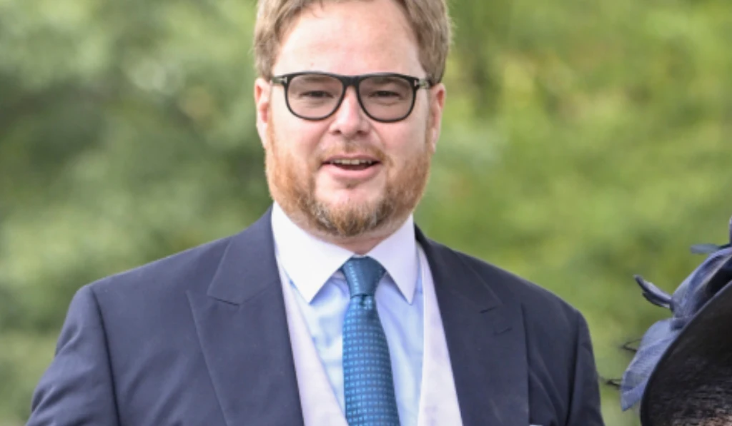 Mężczyzna w okularach, elegancko ubrany w garnitur, białą koszulę i niebieski krawat, uśmiecha się lekko, tło stanowią zielone rozmyte drzewa.