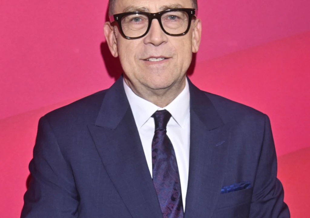 Maciej Orłoś Mężczyzna w średnim wieku w eleganckim garniturze, z krawatem i poszetką, noszący okulary, stojący na tle czerwonej ściany.