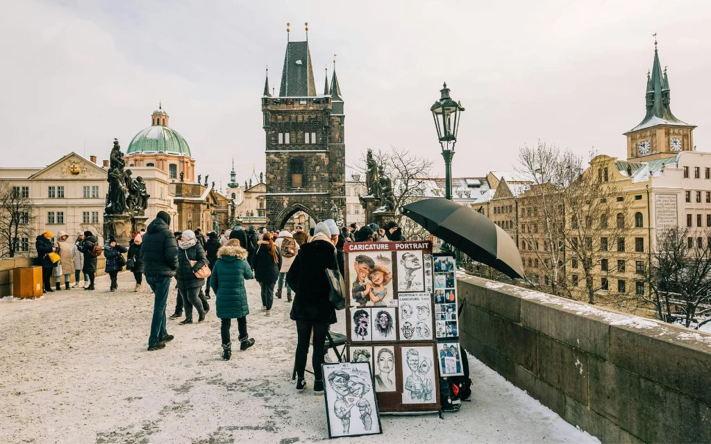 Most ozdobiony historyczną architekturą i licznymi turystami, na pierwszym planie stoisko z karykaturami, wokół przechodnie ubrani na zimowo, w tle wieże i kopuły charakterystyczne dla europejskiego miasta.
