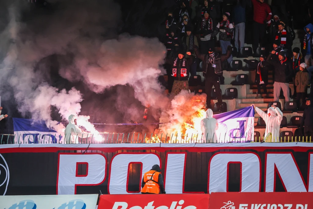 Grupa kibiców na stadionie, wokół unosi się dym z odpalonych rac, widoczny pożar w pobliżu barierki, część osób ubrana w białe kombinezony ochronne, na pierwszym planie baner z napisem Polonia, a w tłumie obecny ochroniarz w pomarańczowej kamizelce.