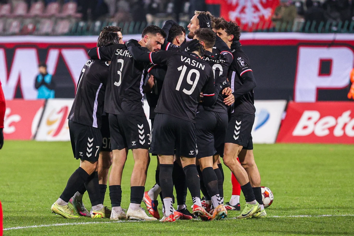 Grupa piłkarzy ubrana w czarne stroje świętuje wspólnie na boisku po zdobyciu gola. Zawodnicy obejmują się, tworząc krąg, wokół widać fragmenty stadionu oraz dużą tablicę reklamową.