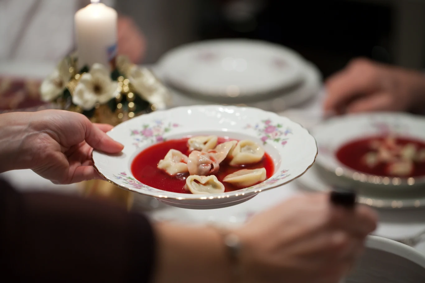 Ręka podaje porcelanowy talerz z czerwonym barszczem i uszkami, w tle widać świąteczną dekorację ze świeczką oraz inne talerze na stole.
