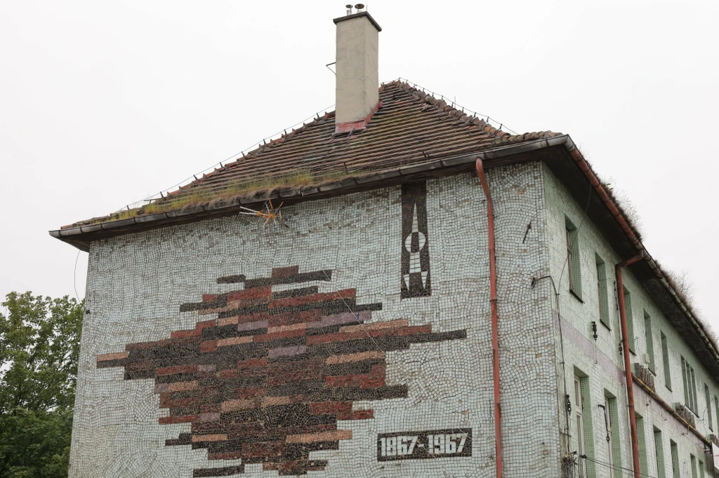 Dawny budynek Huty Szkła Walcowanego w Wałbrzychu Fragment budynku z mozaiką na ścianie bocznej, przedstawiającą ozdobny wzór z napisem oraz datami 1867–1967, dach pokryty dachówką z widocznym kominem i rynnami, w tle drzewa oraz szare niebo.