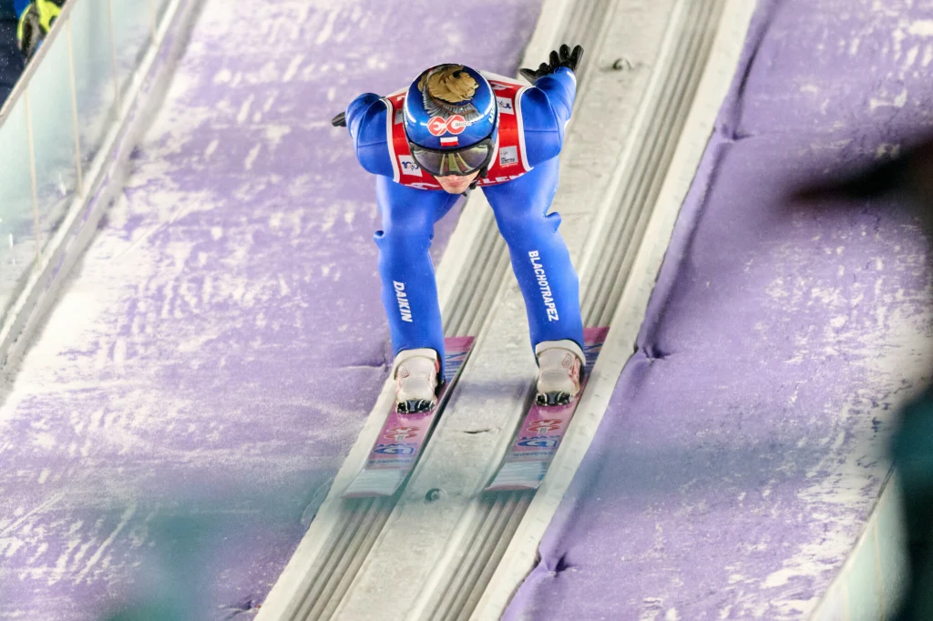 Maciej Kot Skoczek narciarski w niebieskim kombinezonie i kasku, pochylony do przodu, zjeżdża po torze rozbiegowym podczas zawodów na skoczni pokrytej fioletowym śniegiem.