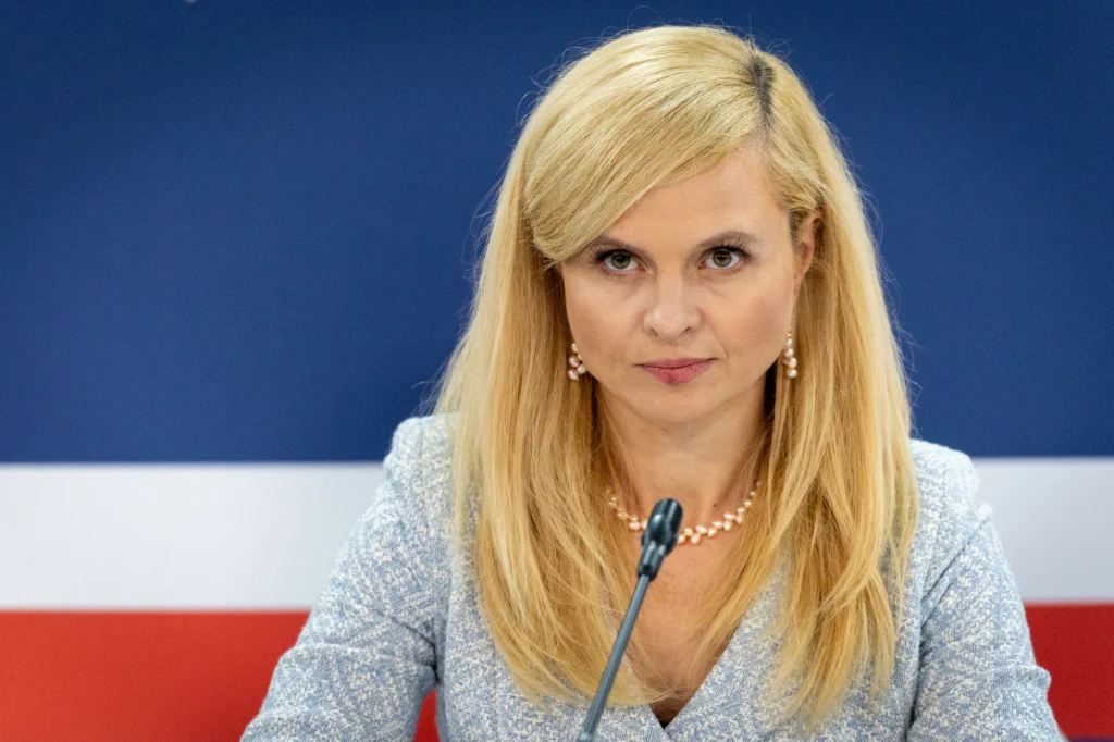 Kobieta o blond włosach w jasnym ubraniu siedząca przed mikrofonem na tle granatowej ściany z biało-czerwoną poziomą linią, patrząca prosto w obiektyw. Ubrana elegancko, nosi perłowy naszyjnik i kolczyki.