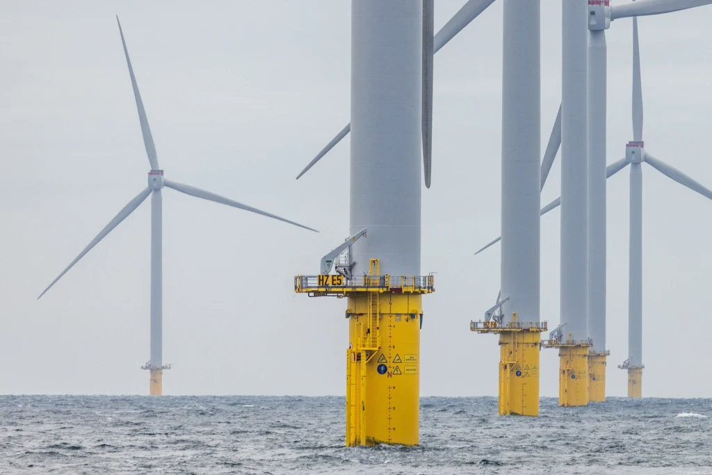 Turbiny wiatrowe ustawione w szeregu na otwartym morzu, z widocznymi żółtymi fundamentami wystającymi ponad powierzchnię wody, szare niebo podkreśla industrialny charakter sceny.